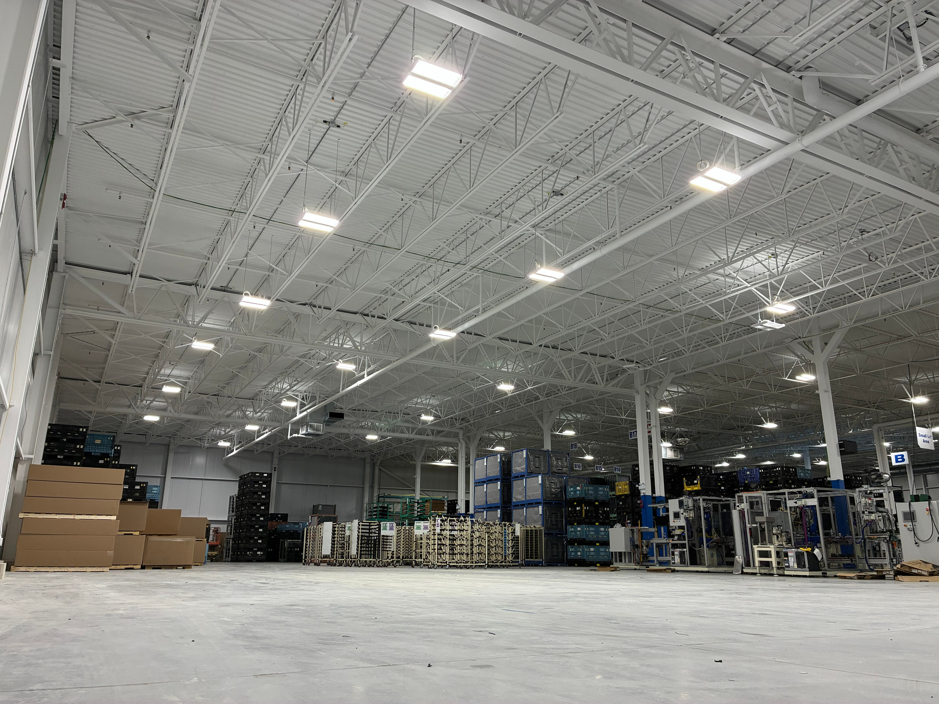 Interior of an industrial factory addition showing steel roof trusses, high-bay LED lighting, and manufacturing equipment